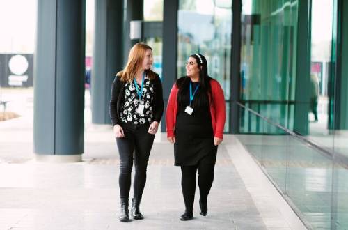 Two women wearing Bupa blue office lanyards and smart casual clothing walk outside a glass office talking together and smiling at each other