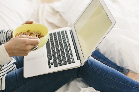 a person with a bowl of cereal and laptop on a bed