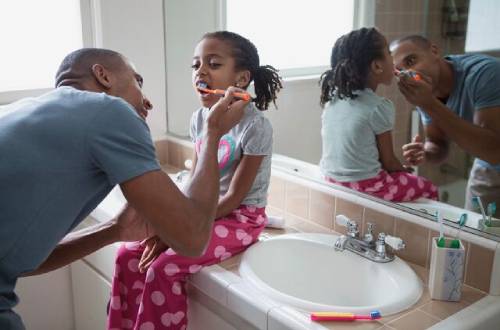 A little girl sits on the side of a basin with her mouth open while a man peers in to her mouth and uses a tooth brush to clean her teeth. The sink next to her does not have the taps running