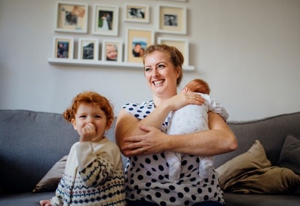 Child sucking thumb, next to mum sat on sofa holding a baby