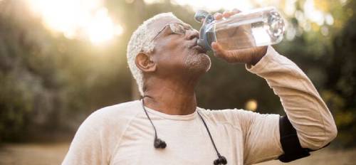 A man stands tipping a clear water bottle up to his mouth so he can drink. His eyes are closed and the sun is settling behind hime