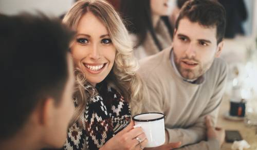 two men and a woman site at a bar  together. The woman is smiling and holding a mug, one man is leaning forward attentively and one man has his back to us and is speaking to the other two people