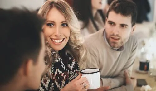 two men and a woman site at a bar together. The woman is smiling and holding a mug, one man is leaning forward attentively and one man has his back to us and is speaking to the other two people