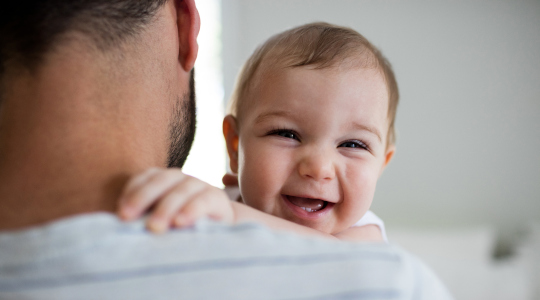 Happy baby smiling at camera over dad’s shoulder