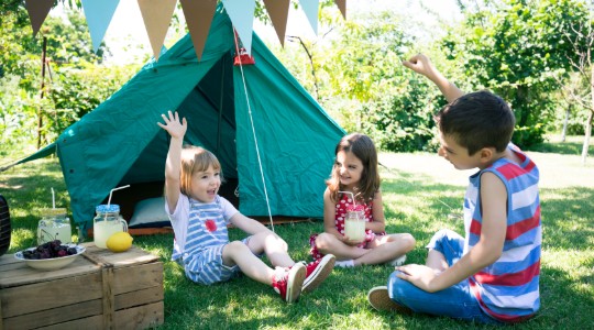 Three children smiling and playing outdoors next to tent