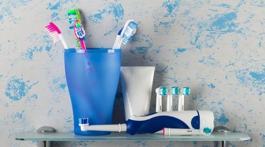 Manual toothbrushes in a pot with electric toothbrush and replacement head laid down on a glass shelf