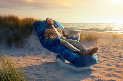 A man sits on a fluffy blue dentist chair on the beach, smiling and looking relaxed