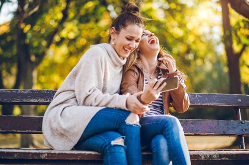 Two girls sitting on a bench, laughing while taking a selfie.