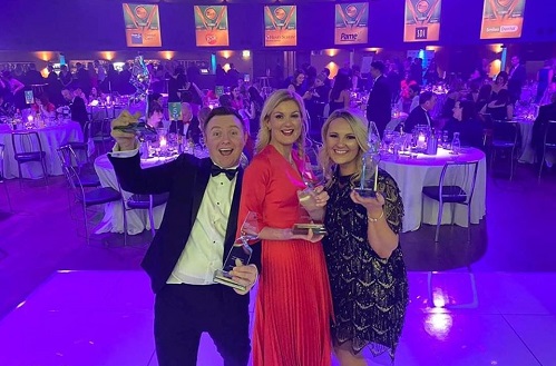 Photo of two women and one man at awards ceremony holding up awards and smiling